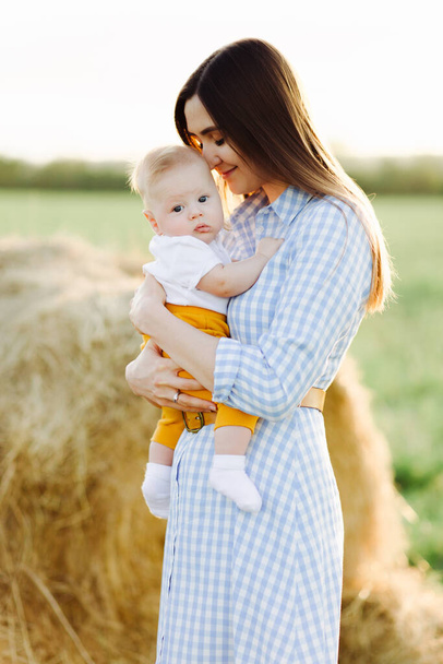 una joven madre feliz con un niño pequeño en sus brazos, suavemente apoyado contra su cabeza, se encuentra en medio de un campo cerca de un pajar recogido en los rayos de la puesta del sol
 - Foto, imagen