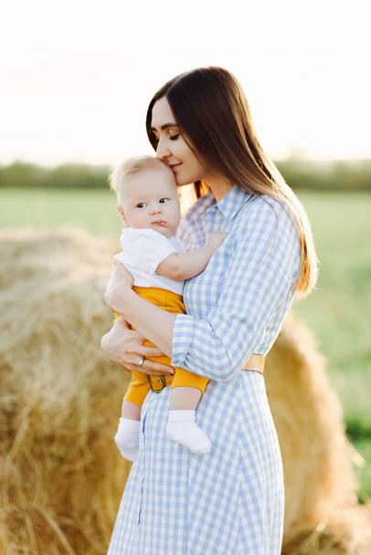 幼い子供を抱えた幸せな母親が優しく頭をキスして夕日の光の中の畑の真ん中に立っています - 写真・画像