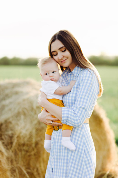een jonge gelukkige moeder met een klein kind in haar armen, zachtjes leunend tegen zijn hoofd, staat in het midden van een veld in de buurt van een verzamelde hooiberg in de stralen van de zonsondergang - Foto, afbeelding