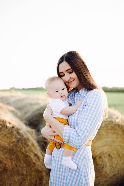 uma jovem mãe feliz com uma criança pequena em seus braços, gentilmente encostada à cabeça, fica no meio de um campo perto de um palheiro coletado nos raios do pôr do sol
 - Foto, Imagem