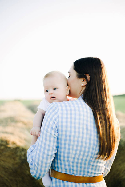 una joven madre con un vestido azul a cuadros se para con la espalda a un niño pequeño mirando a la cámara sobre su hombro, en los rayos del sol poniente de verano
 - Foto, Imagen
