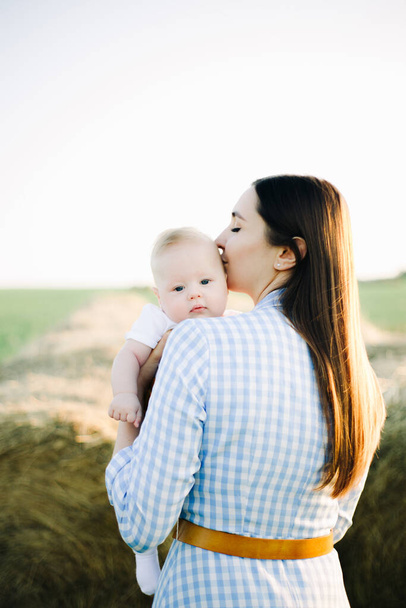 a young mother in a blue checked dress stands with her back to a small child looking at the camera over her shoulder, in the rays of the setting summer sun - Foto, imagen