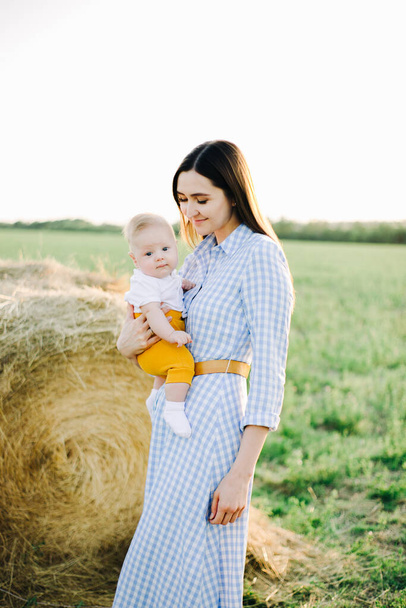 uma mãe bonita nova com uma criança pequena em seus braços está em um vestido, no meio de um campo perto de um palheiro coletado nos raios do por do sol dourado
 - Foto, Imagem