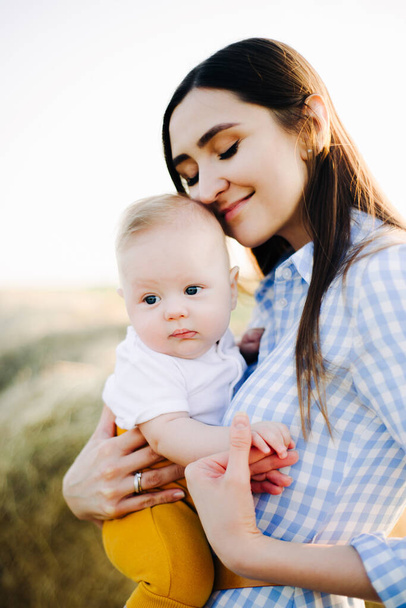 молодая счастливая мать с маленьким ребенком на руках, нежно прислонившись к его голове, стоит посреди поля рядом с собранным стогом сена в лучах заката
 - Фото, изображение