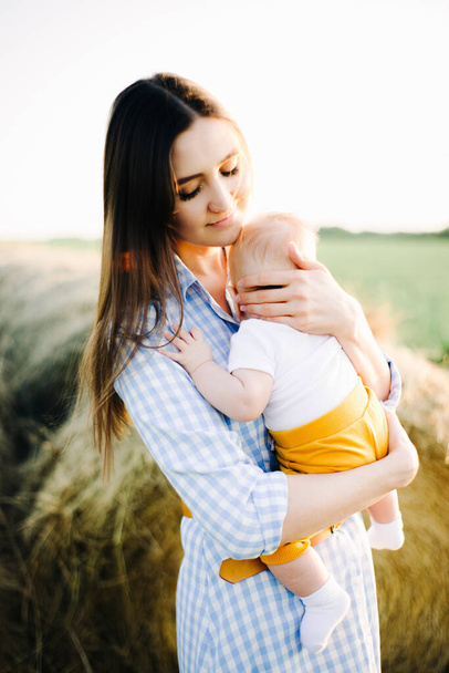 Kollarında küçük bir çocukla genç ve güzel bir anne, altın gün batımının ışınlarında toplanmış samanlığın yanındaki bir tarlanın ortasında elbise içinde dikiliyor. - Fotoğraf, Görsel