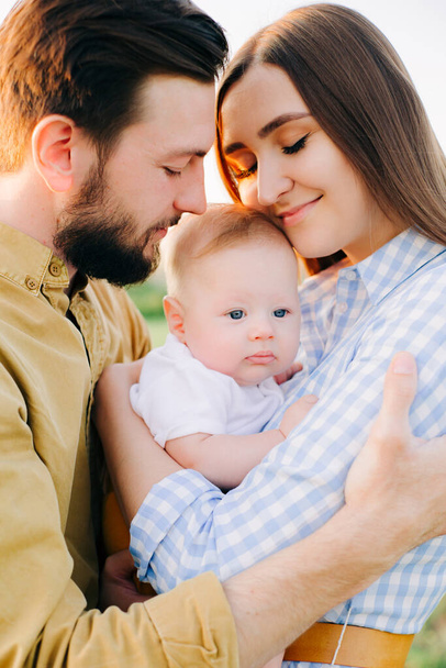 portrait of a happy young family, where mom and dad hug each other, holding their little child between them in the rays of the sunset, sun - 写真・画像