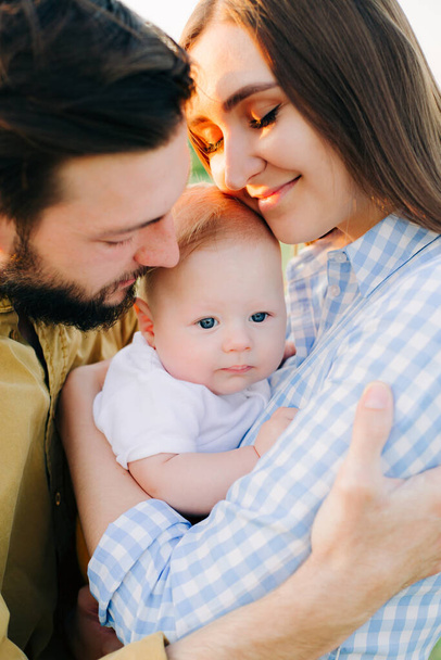 портрет счастливой молодой семьи, где мама и папа обнимают друг друга, держа своего маленького ребенка между ними в лучах заката, солнца
 - Фото, изображение