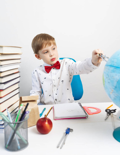 Child education: a little boy in a shirt and a red bow-tie sits at a desk and measures distances on the globe with a compass. Portrait. - Fotoğraf, Görsel