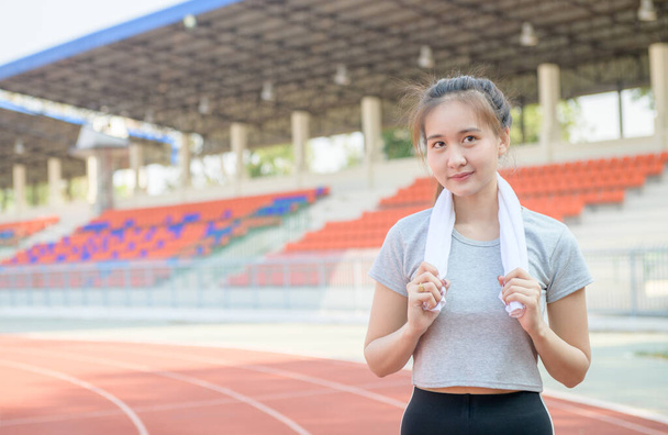 Młoda Azjatka z białym ręcznikiem na szyi, stojąca na stadionie. Koncepcja sportu i ćwiczeń. - Zdjęcie, obraz