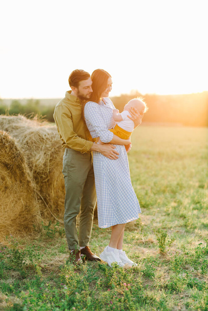 happy young family, mom and dad standing in a field near a haystack, holding hands, father holding a small happy child in the rays of the sunset sun - Photo, Image