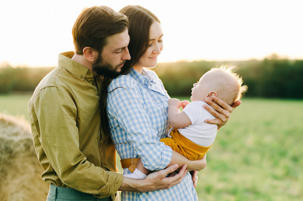 retrato de una familia joven y feliz, donde mamá y papá se abrazan, sosteniendo a su pequeño hijo entre ellos en los rayos de la puesta del sol, el sol
 - Foto, Imagen