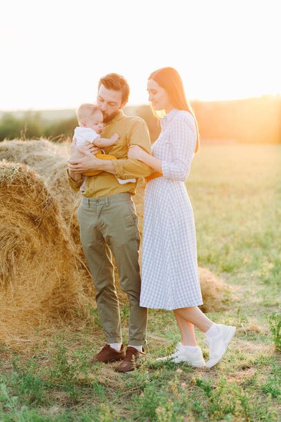 happy young family, mom and dad standing in a field near a haystack, holding hands, father holding a small happy child in the rays of the sunset sun - Photo, image