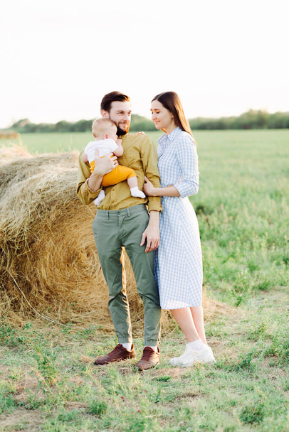 happy young family, mom and dad standing in a field near a haystack, holding hands, father holding a small happy child in the rays of the sunset sun - Fotografie, Obrázek