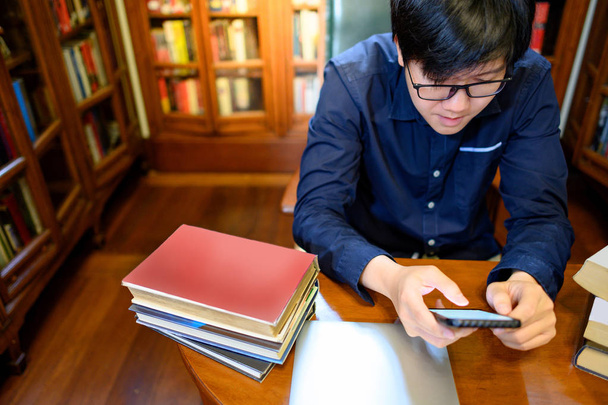 Asiatique homme étudiant à l'université en utilisant smartphone et livre de lecture à proximité bibliothèque vintage ou bibliothèque dans la bibliothèque du collège. Ressources pédagogiques pour la recherche en éducation. Concept d'opportunité de bourse
 - Photo, image