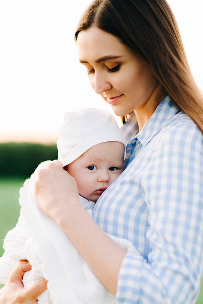 portrait of a young mother in a blue checked dress, holding a small child to her chest, in the rays of the setting sun - Foto, Imagen