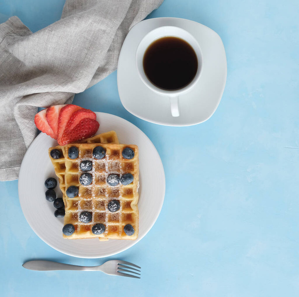 Viennese or Belgian waffles on a white plate with fresh organic berries in icing sugar. Nearby is a cup of fresh coffee, a fork and a table napkin. Place for text. Blue background - Photo, Image