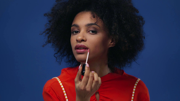riccio africano americano ragazza applicando lucidalabbra isolato su blu
 - Filmati, video