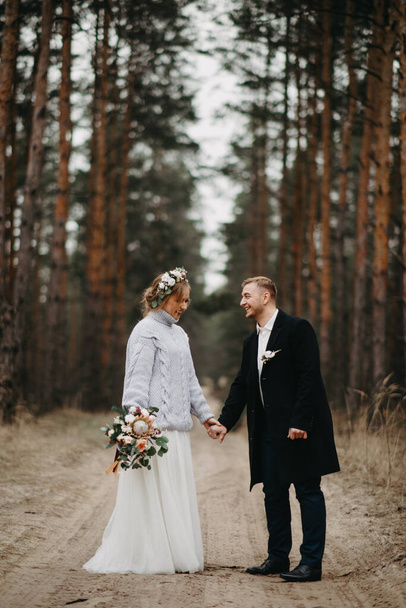 Los recién casados se toman de la mano y caminan por un sendero forestal en un bosque de coníferas entre pinos
. - Foto, Imagen