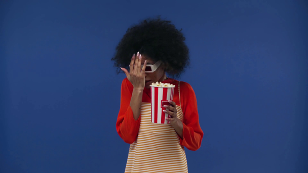 Afrikai amerikai lány 3D-s szemüvegben popcornt eszik, elszigetelve a kéktől. - Felvétel, videó