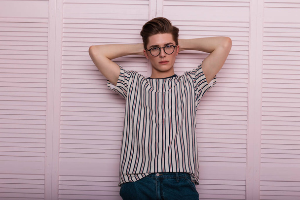 Europea bastante elegante joven hipster hombre en gafas de moda con peinado de moda en una camiseta de rayas vintage posando cerca de una pared de madera de color rosa en la habitación. chico guapo modelo de moda posando en el interior
 - Foto, imagen