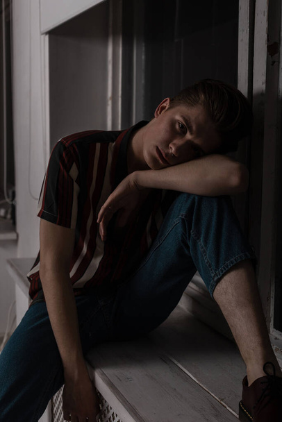 Pretty stylish young man in a striped vintage shirt in blue jeans is resting near the window in the room in the evening. European guy model in trendy clothes enjoys relaxing indoors. Youth menswear. - 写真・画像