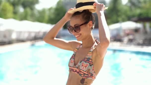 hezká mladá žena v bikinách, klobouk a sluneční brýle radostně se usmívá na pozadí bazénu. - Záběry, video