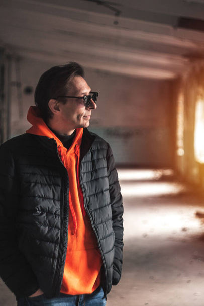 guy in a dark jacket and orange sweatshirt posing against the backdrop of warm light from the windows - Fotografie, Obrázek