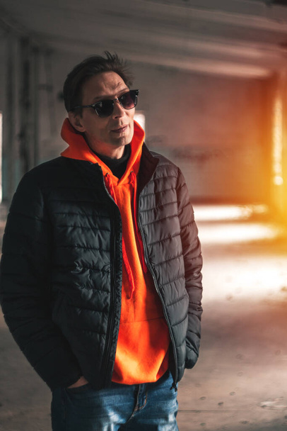 guy in a dark jacket and orange sweatshirt posing against the backdrop of warm light from the windows - Фото, изображение