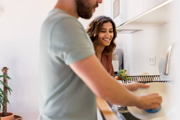 Paar teilt sich Hausarbeit. Glückliche Frau beobachtet Mann beim Geschirrspülen.  - Foto, Bild