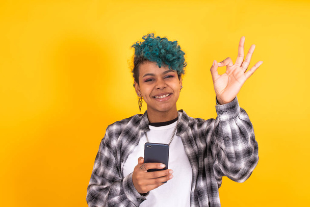jong brunette meisje met mobiele telefoon en oke teken geïsoleerd op gele achtergrond - Foto, afbeelding