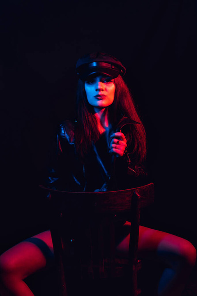 fashionable portrait of a young sexy girl in a leather jacket and cap with a neon light - Фото, изображение
