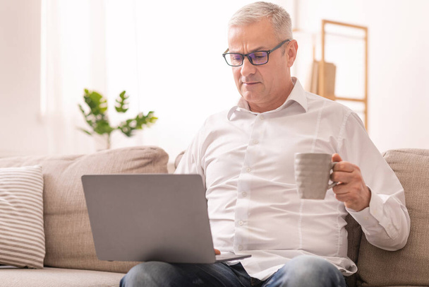 Oudere man die laptop gebruikt en thee drinkt - Foto, afbeelding