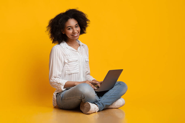 Afro donna seduta sul pavimento, utilizzando il computer portatile e sorridendo alla fotocamera
 - Foto, immagini