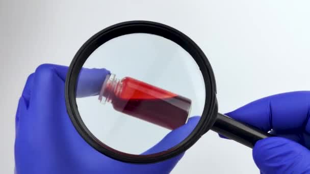 Amostra de sangue para análise. A mão dos médicos detém uma lupa em que um tubo de teste com uma amostra de sangue para pesquisa em laboratório. Lugar para texto
 - Filmagem, Vídeo