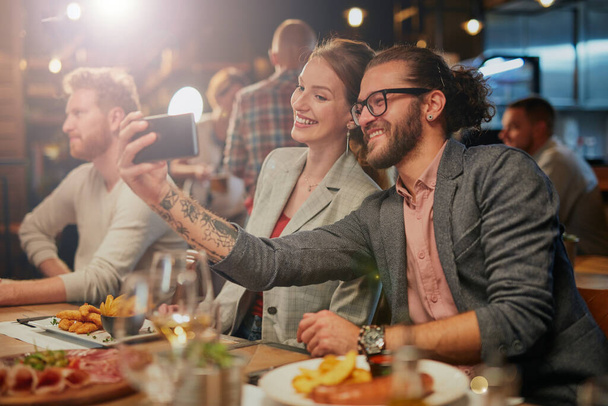 Cute caucasian fashionable couple sitting in restaurant at dinner and taking selfie. - 写真・画像
