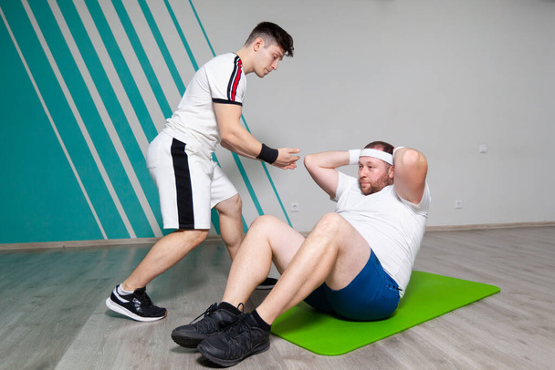 Fat man looks exhausted while doing exercises on abdominal muscles lying on the mat in the fitness gym under strict control of personal instructor. Overweight - Photo, image