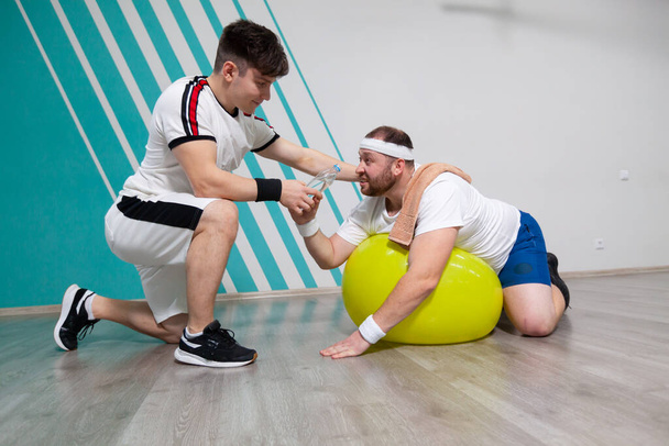 Overweight man is lying on the fitness ball in the gym while his personal trainer offers him a bottle of water. Fat man is dying of thirst and looking exhausted after a hard training in fitness group - 写真・画像