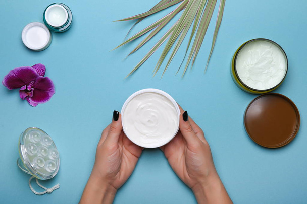 Moisturizing cream in woman's hands over blue table with cosmetic jars flat lay - Foto, immagini
