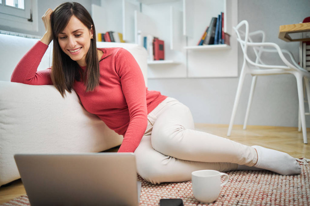 Young attractive smiling woman sitting at home on the floor and using laptop. - Foto, Bild