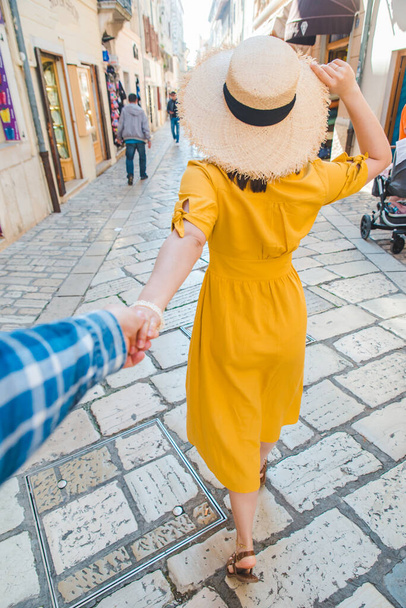 sledovat můj koncept. žena ve žlutých slunečních šatech v slaměném klobouku kráčející kupředu u malého letoviska City Street držící muže za ruku. letní dovolená - Fotografie, Obrázek