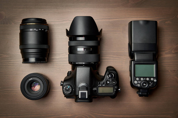 top view of modern digital camera equipment - DSLR with attached zoom lens and hood, lenses and external flashlight on wooden table - Photo, Image