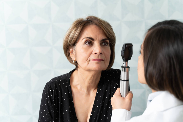 Gériatre femelle vérifiant l'œil de la femme âgée avec otoscope en clinique
 - Photo, image