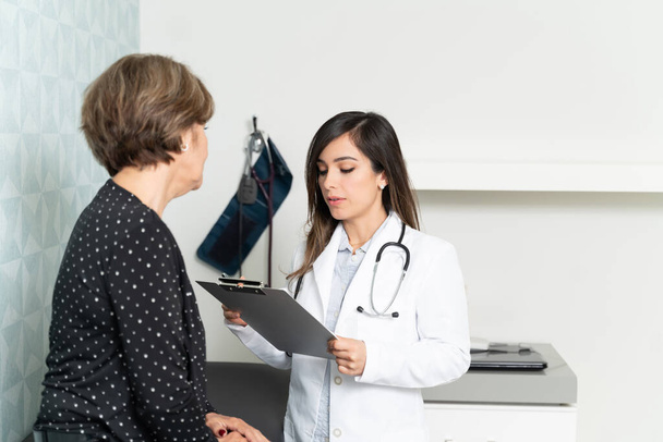 Vrouwelijke geriater die medisch dossier onderzoekt terwijl oudere patiënt in kliniek zit - Foto, afbeelding