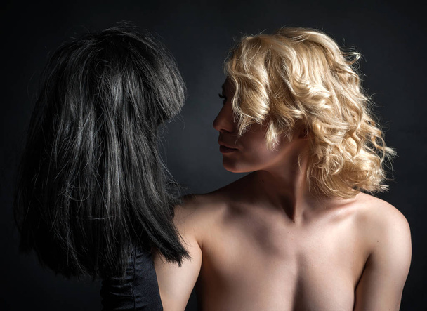eine junge blonde Frau mit Perücke, die eine schwarze Perücke in der Hand hält. auf schwarzem Hintergrund - Foto, Bild