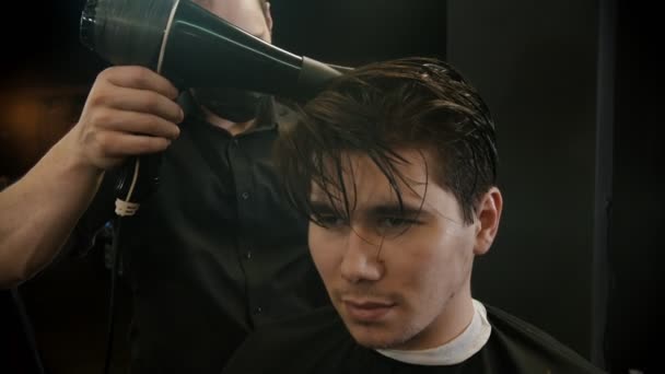 Barber in black mask styling his client hair with hairdryer and brush - Filmati, video