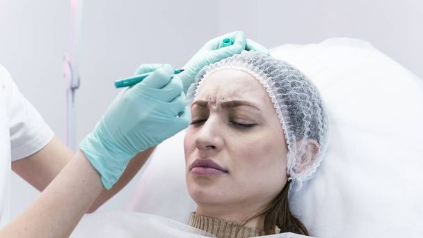 The young beautician doctor preparing to making injection in female forehead. The doctor cosmetologist makes anti-aging treatment and face lift procedure. Concept of beauty salon and plastic surgery clinic. - Фото, зображення