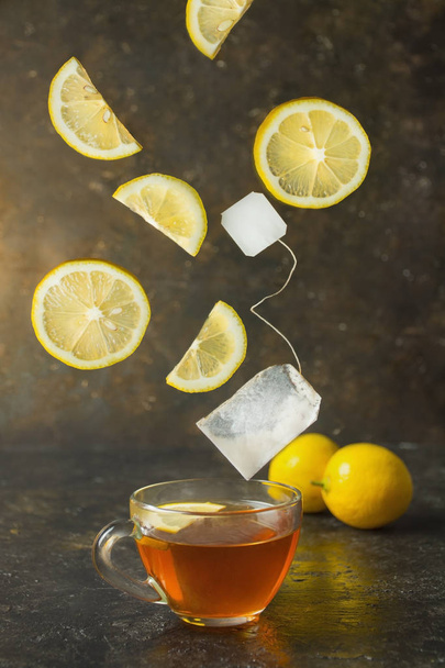 Té con limón en una taza de vidrio sobre un fondo oscuro. Los ingredientes caen en el té. El efecto de la levitación. Comida en movimiento
. - Foto, Imagen