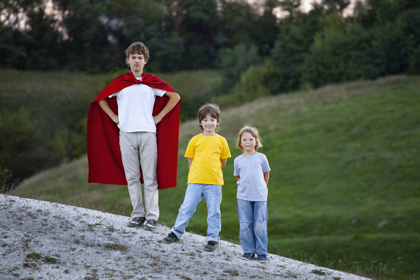 Boys playing superheroes outdoors, teenage superhero in a red cloak on a hill - Фото, изображение
