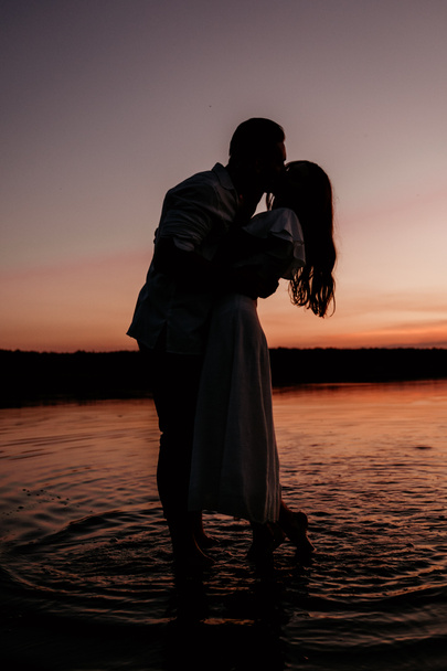 Young couple embracing and kissing in the water on Sunset. Two silhouettes against the sun. Romantic love story. - 写真・画像