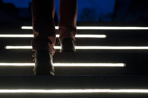 Männliche Beine in Stiefeln erklimmen in einer Winternacht die beleuchteten Steintreppen. Ansicht von unten. - Foto, Bild